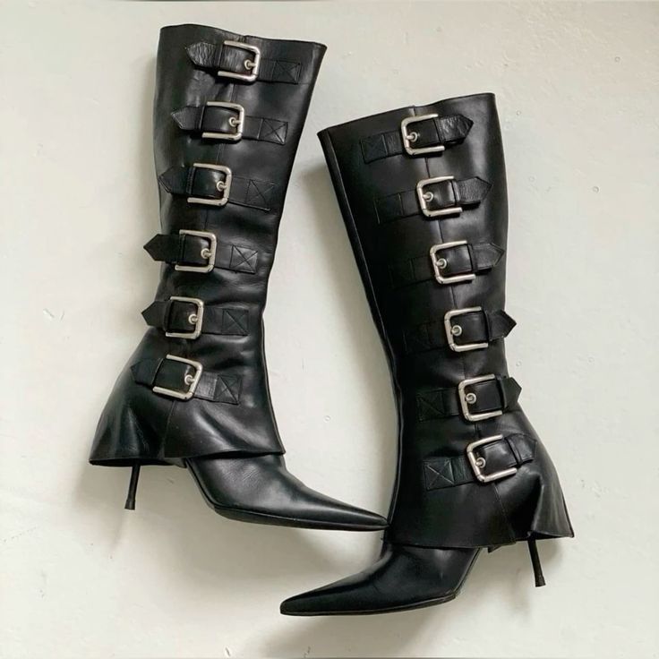 a pair of black leather kneehigh boots with multiple silver buckles is displayed against a white background.  the boots have pointed toes and stiletto heels. one boot is positioned slightly behind the other, showcasing both boots' full profiles.  the boots appear to be vintage or retro in style.