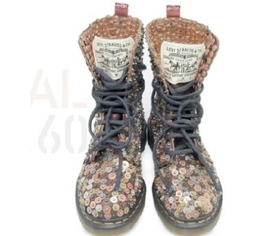 the image shows a pair of dr. martens boots that have been intricately embellished with a multitude of small buttons, coins, and other metal pieces.  the boots are dark brown/black leather and feature vintage levi's labels sewn onto the tongues. the embellishments are densely packed, creating a textured and visually striking surface.  the boots are positioned slightly apart against a plain white background. the overall impression is one of handmade, artistic, and highly individualized footwear.