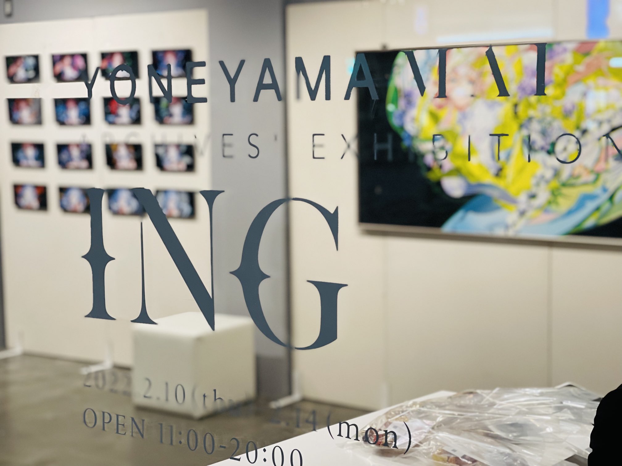 a view through a glass door into an art gallery with an exhibition titled "yoneyamantal's exhibition "ing"". the gallery is brightly lit and features several paintings on the walls. the exhibition runs from february 10th to 14th, with daily opening hours from 11:00 to 20:00.