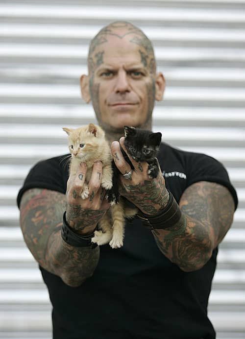a man with heavily tattooed arms is holding two kittens in his hands. one kitten is white and orange, the other is black. the man is wearing a black shirt and has short, black hair. he is looking at the camera with a serious expression. the background is a gray, textured wall.