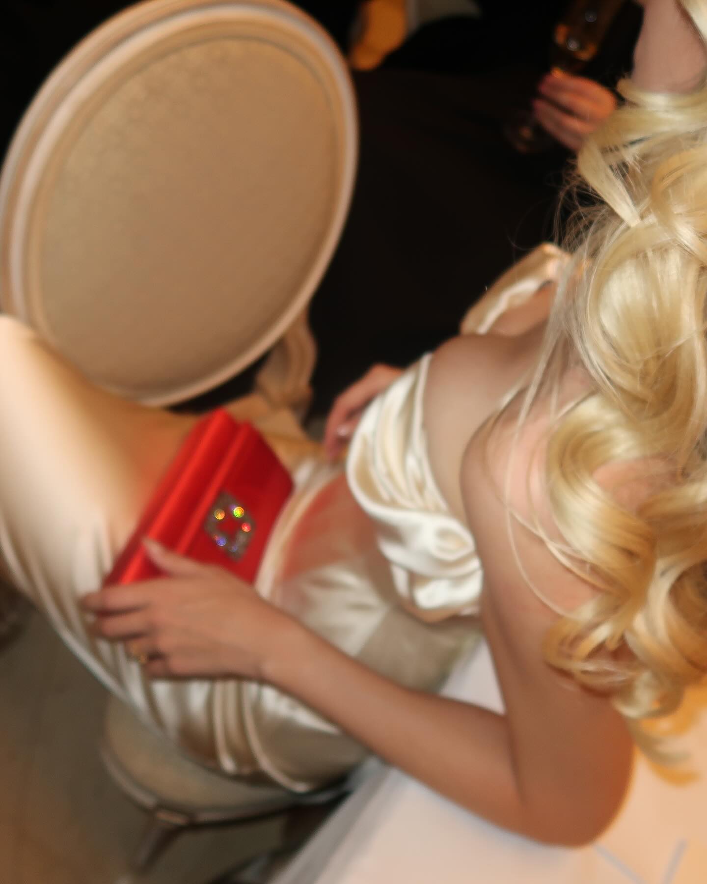 a blonde woman in a white dress with a red clutch bag. she is looking off to the left. there is a chair and a table behind her. the woman is wearing a sleek dress that emphasizes her figure. her hair is styled in a loose wave and looks soft and flowing. the red clutch bag adds a pop of color and contrasts beautifully with the white dress. the woman's elegant and glamorous look is perfect for a night out or a special event. the image is wellcomposed and the lighting is soft and flattering.