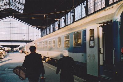 an eyelevel, full shot depicts a vintage photograph of a train station's interior. a train is parked on the platform, its doors open, and two figures stand nearby, one carrying luggage.  the train is a blue and white passenger train, with numerous windows and doors. the station's architecture is visible, showcasing a highceilinged structure with glass roofs and metal frameworks. the overall mood is one of travel and departure, evoking a sense of nostalgia.  the photo appears to be taken with an older camera; the colors are slightly muted, and the image has a grainy texture.