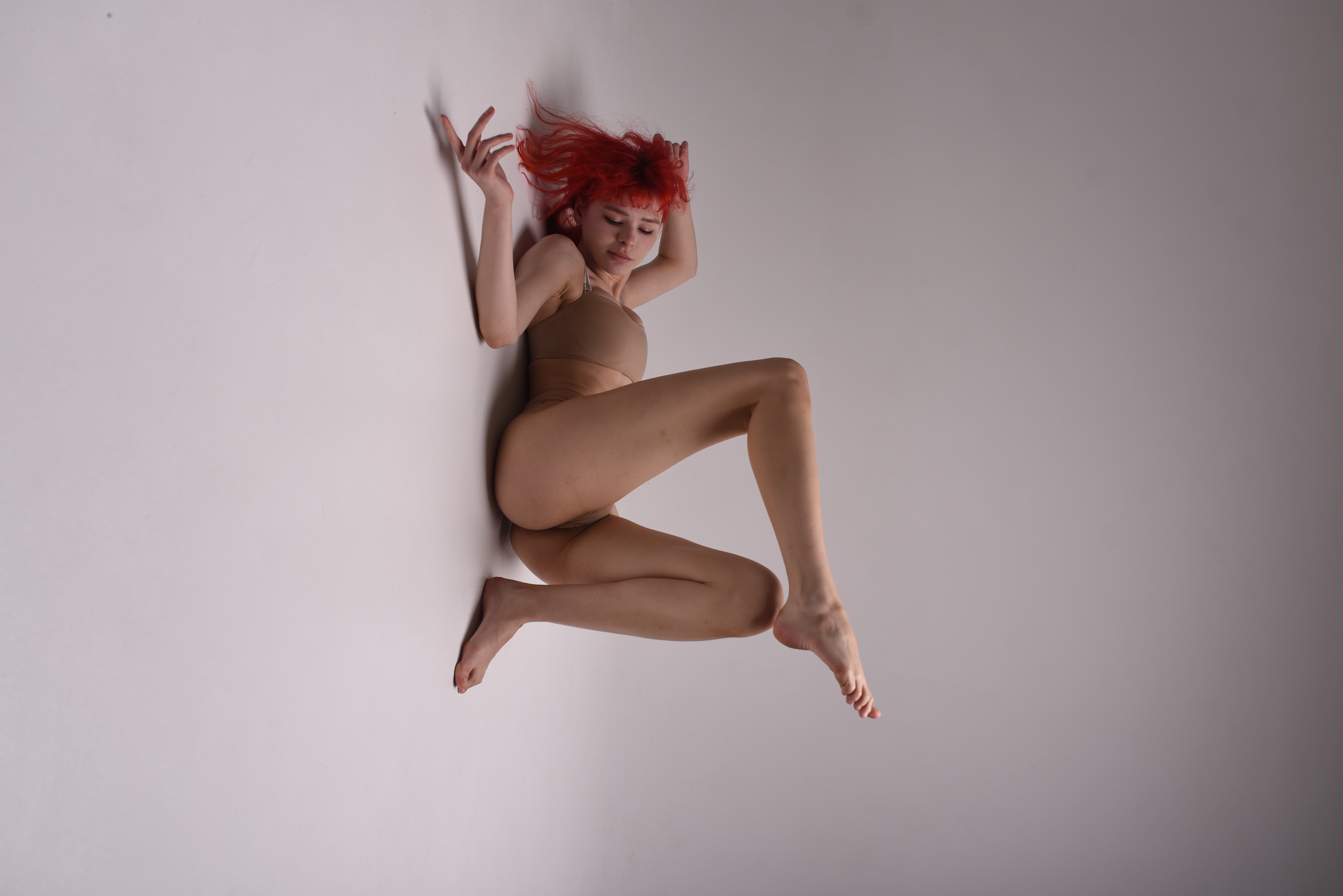 a woman with red hair, wearing a nude bodysuit, is captured middance move in a studio setting. the soft lighting emphasizes her graceful pose and the flexibility of her body.  the image evokes a sense of modern, contemporary dance.