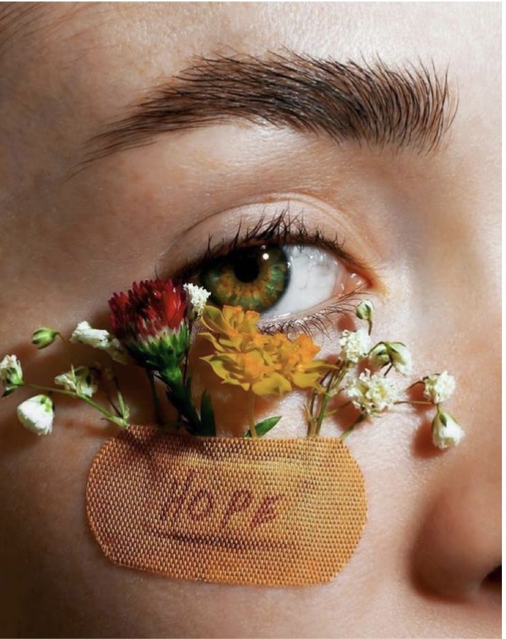 a closeup shot of a woman's face, specifically focusing on her eye.  a small, floral arrangement of various wildflowers (red, yellow, and white) is delicately placed over her eye, partially covering it, and resting on a small, beige bandage with the word "hope" written on it. the woman's makeup is minimal, and the overall aesthetic is delicate and artistic. the image has a soft, dreamy quality, emphasizing the beauty of the flowers and the hope conveyed by the message on the bandage.