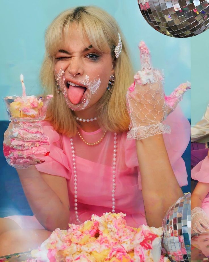 a closeup shot of a young blonde woman wearing a pink dress and white gloves, playfully covered in cake frosting. she's winking at the camera with her tongue sticking out, holding a small piece of cake in one hand and another cake is situated in front of her. there are disco balls in the background, contributing to a fun and celebratory atmosphere. the overall style is reminiscent of 80s aesthetics, bright and cheerful.