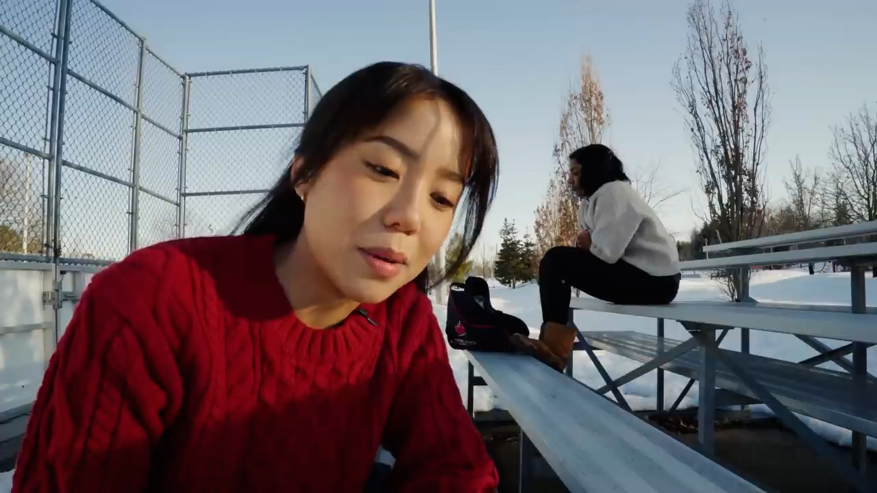a selfiestyle photo taken by a young woman wearing a red sweater. she is sitting on bleachers with ice skates next to her. another young woman is in the background, out of focus. the bleachers are covered in snow and the ground is also snowcovered. the photo appears to be taken on a sunny winter day.