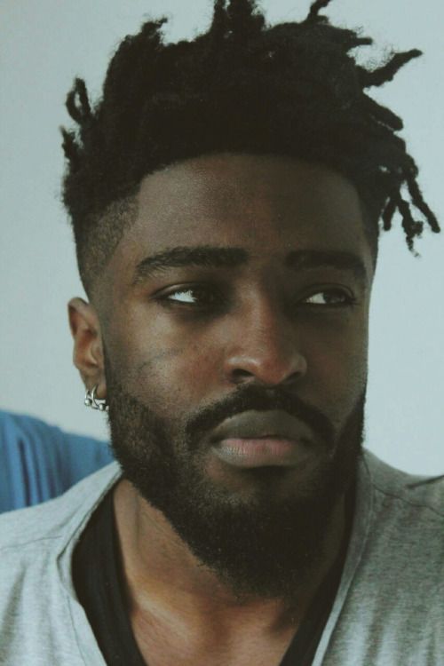 a closeup portrait of a young black man with dreadlocks and a beard. he is looking directly at the camera with a serious expression. he is wearing a gray tshirt. the image is focused on his face.