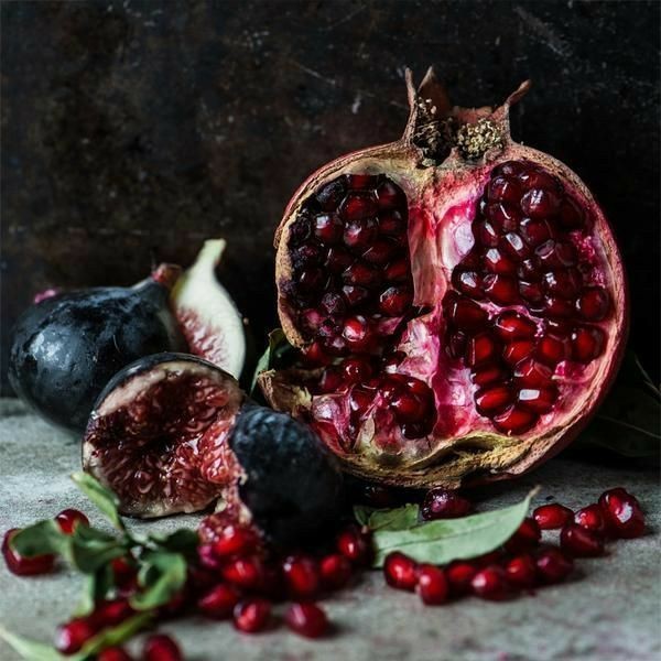 a close up still life photograph of a halved pomegranate with its seeds exposed, next to a whole and halved dark purple fig. scattered around the fruits are red pomegranate seeds on a rustic dark textured background. the image has a moody and slightly dark aesthetic.