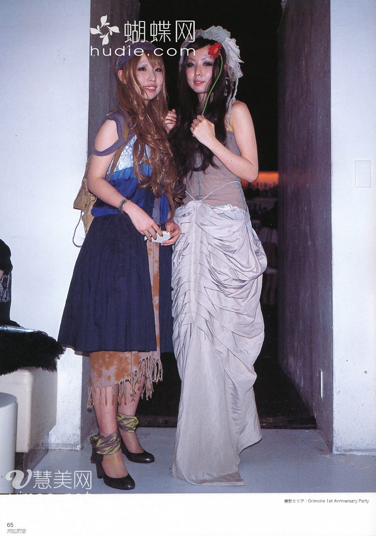 two young women pose for a photo in an interior setting. the woman on the left wears a blue, white, and brown dress with a bohemian feel. the skirt features a layered design, and she pairs the look with strappy sandals. the woman on the right wears a more dramatic outfit, a long, flowing, layered, and ruffled dress in a light, muted tone. she holds a single red flower and wears a matching floral embellishment in her hair. the background suggests a party or social gathering, with glimpses of furniture and another room visible.  kw: fashion, style, japanese fashion, party, friends, social event, portrait, dresses, bohemian, layered clothing, ruffles, indoor photography