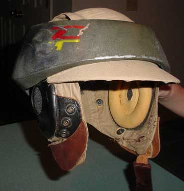 a close up of a vintage military flight helmet. the helmet is beige with brown leather ear flaps. the top of the helmet is covered in green fabric with a black and red logo. the helmet has a large, yellow foam pad on the right side. the helmet is sitting on a table.