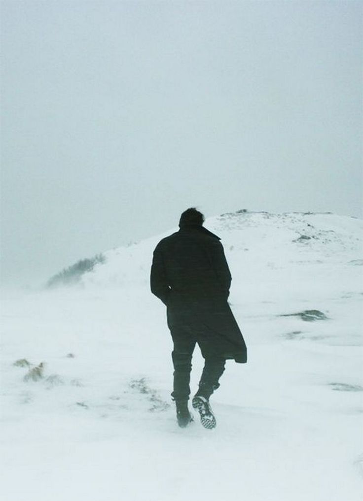 the image shows a lone man walking away from the camera across a vast, snowcovered landscape.  the sky is a muted greywhite, and the overall atmosphere is cold, bleak, and lonely. the man is dressed in a dark coat and boots, and his silhouette is stark against the snow.  the scene is minimalist and emphasizes the vastness of the winter landscape and the isolation of the figure.  the overall mood is quiet and contemplative.