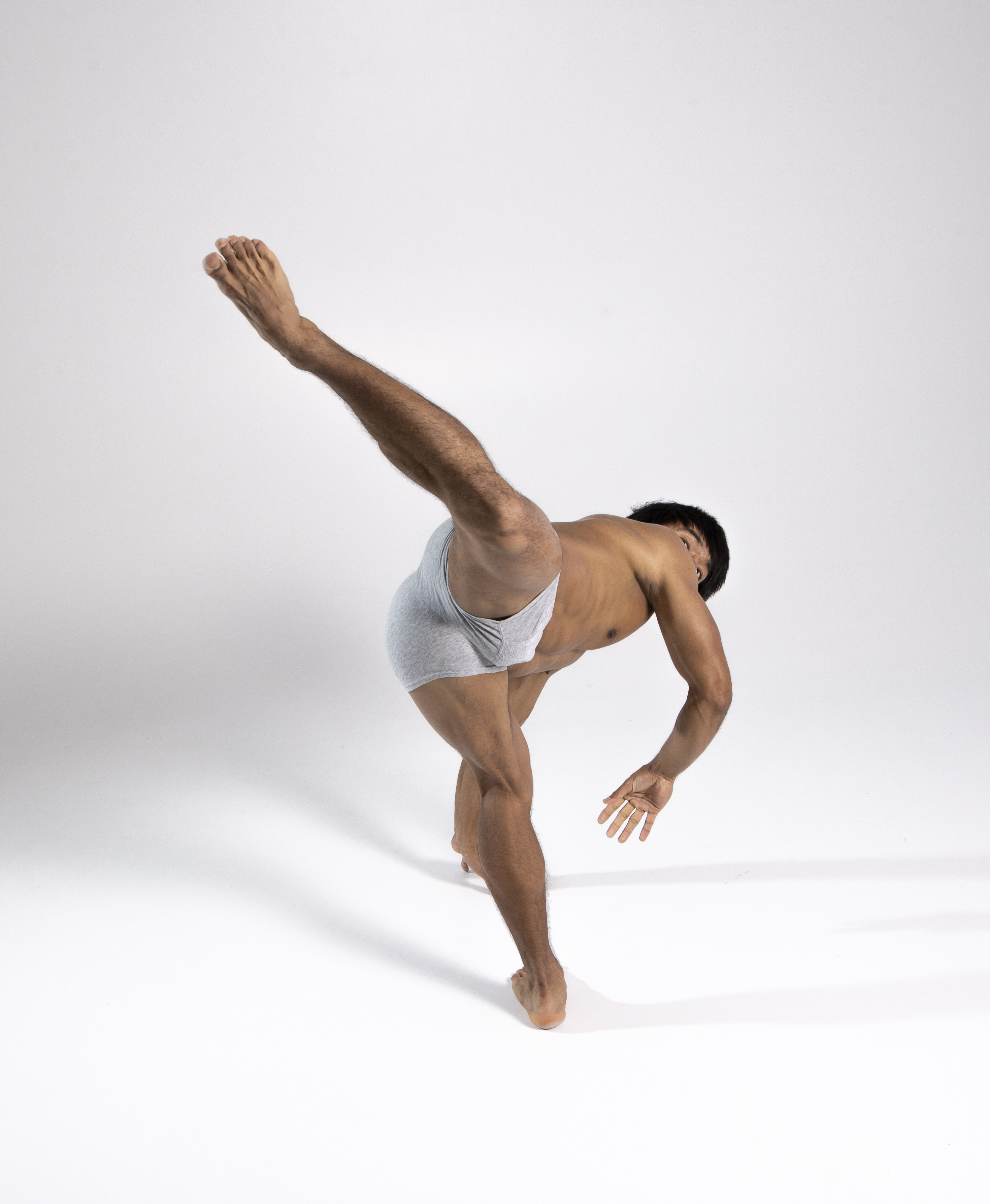 a fulllength studio shot of a muscular, athletic man in gray underwear is captured midpose. he is barefoot and positioned against a plain white background. his body is contorted in a dancelike position, balancing on one leg while the other is raised high in the air. his arms are extended for balance. the lighting emphasizes his muscular physique and dynamic movement. the focus is on his body and the expression of movement, highlighting his physical fitness and flexibility.