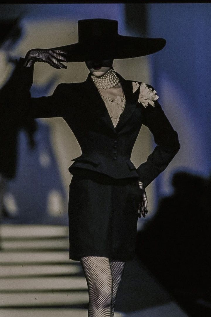 a black and white photo of a runway model in a high fashion black suit. the model is wearing a widebrimmed black hat, long black gloves, and fishnet stockings. the suit is cinched at the waist and has a peplum detail. the model is wearing a pearl necklace and a small white flower is pinned to her jacket. the background is dark and moody, and the stairs in the background add depth to the image. the overall feel is elegant, chic, and slightly gothic.