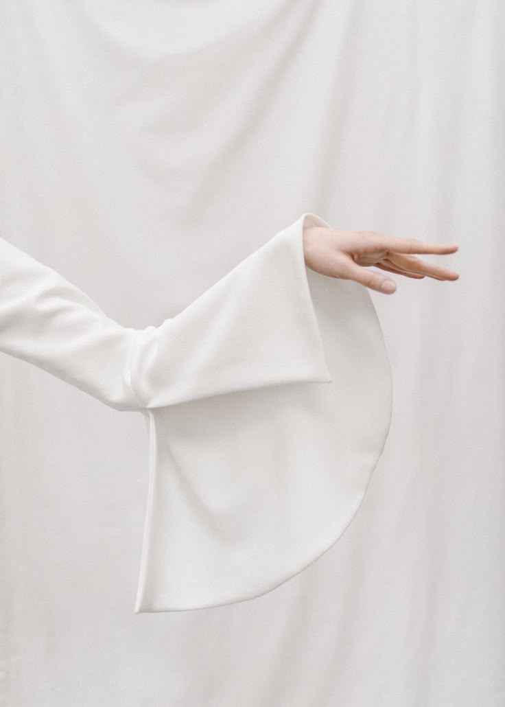 a closeup shot shows a white, longsleeved garment with a flared sleeve that extends towards the right side of the frame. a hand emerges from the sleeve, with the fingers extended, positioned in front of a backdrop of more white fabric. the entire composition is a study in white tones.