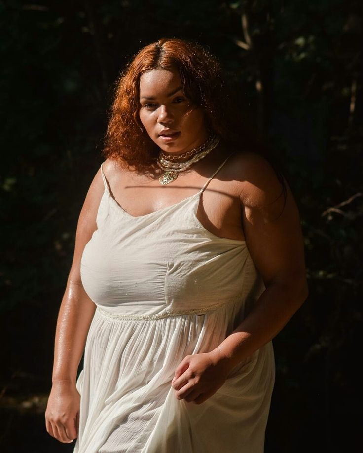 a young woman with red curly hair is posing for the camera in a beautiful white dress, standing outside in a green and leafy forest. the sun is shining through the trees, casting a golden glow on the model's face and body. she is smiling, wearing a necklace with a golden pendant, and her beauty is enhanced by her natural surroundings. the image is a testament to the power of nature and the beauty of femininity.