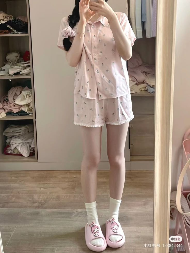 a person is standing in front of a mirror, wearing a pink pajama set with a pattern. the set includes a shortsleeved collared shirt and shorts with a ruffled edge. the person is wearing white socks and pink my melody slippers. the reflection shows a closet or wardrobe behind the person. there is a hair tie on their wrist. the scene is indoors.