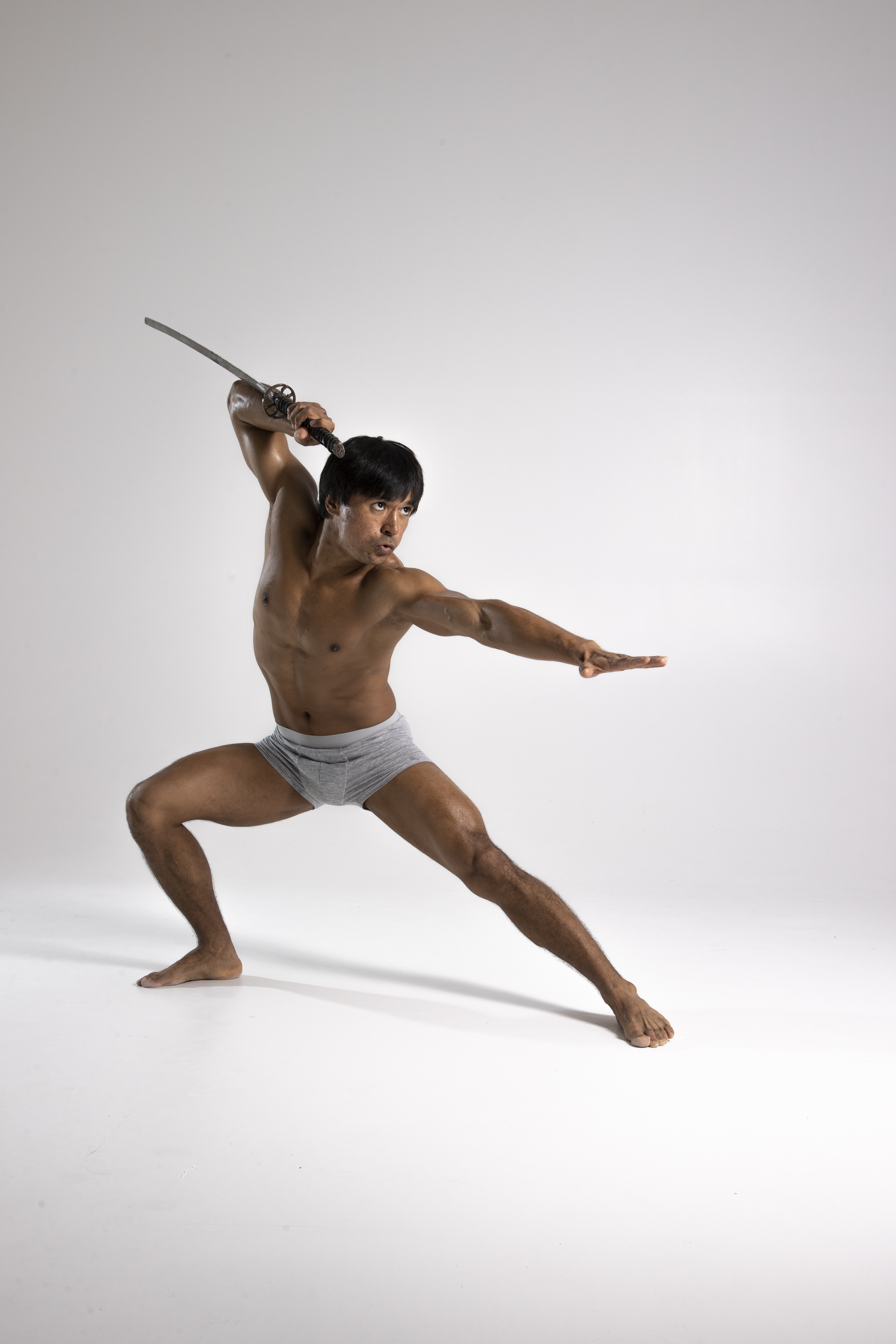 a shirtless asian man in gray underwear stands barefoot with his legs apart and bent as if in a martial arts stance. he's holding a katana sword above his head and extending his other arm straight out to the side. he looks ready to strike, with a determined expression. the background is a plain white or light gray, making the man the primary subject of the image.
