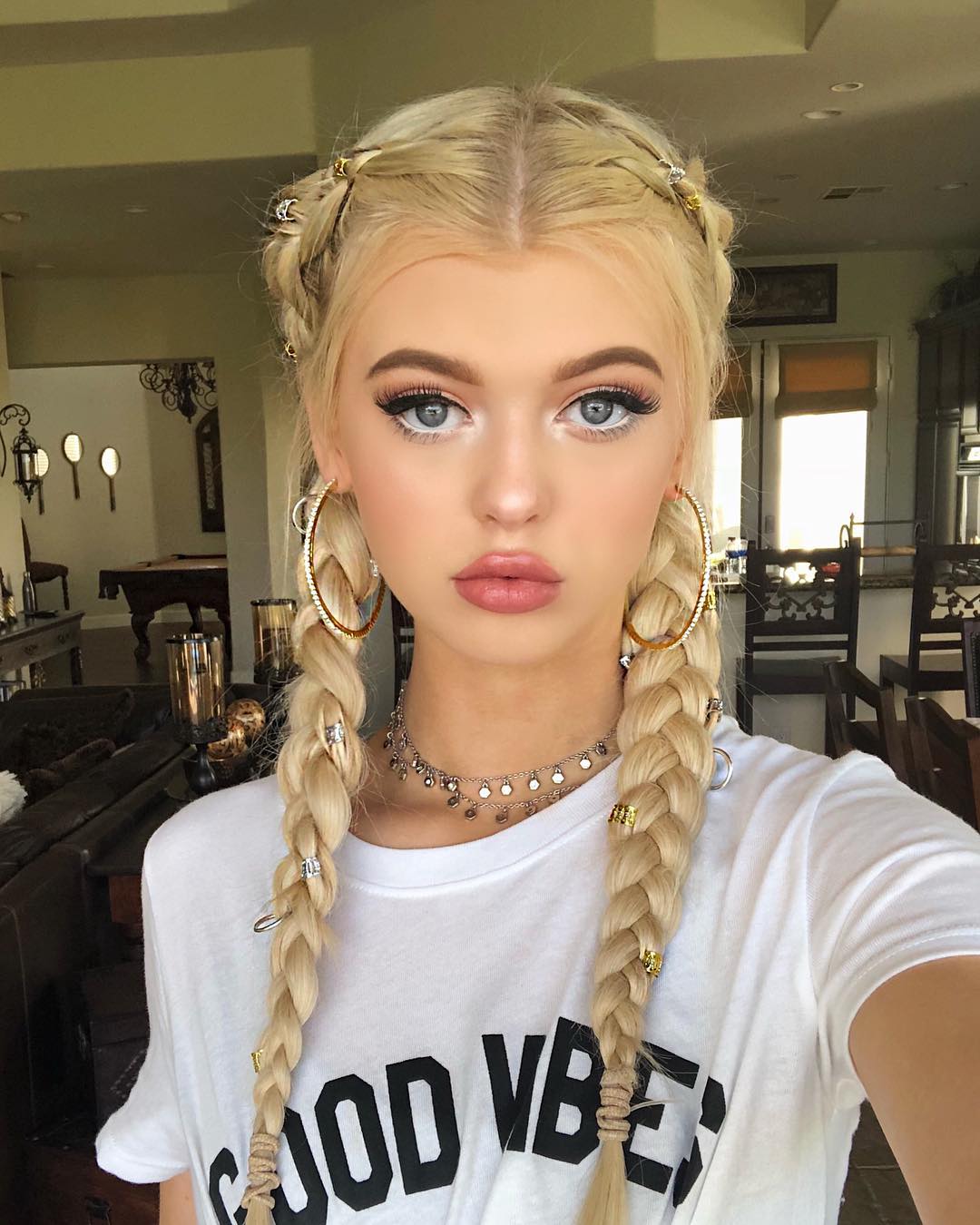 a woman with blonde hair styled in braids is wearing hoop earrings, a necklace, and a white tshirt that says "good vibes." she has blue eyes and is wearing makeup. her hair is styled in pigtails with decorative accessories throughout the braids.