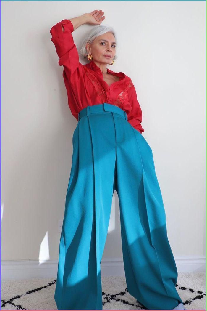 a woman with grey hair stands with her arms raised and a wide smile on her face. she is wearing a bright red shirt and highwaisted blue wide leg pants.
