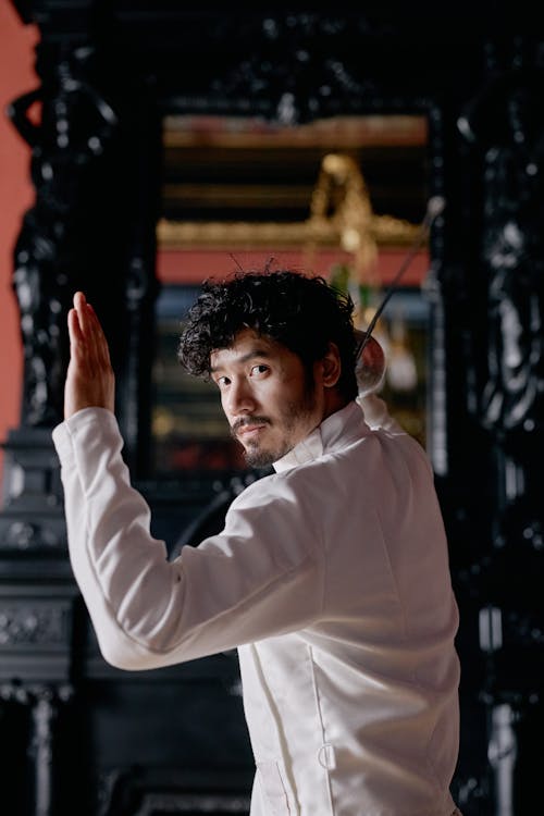 a portrait of a man in a white fencing jacket, posing with a foil in his hand. he is standing in an elegant, dark indoor setting, against a backdrop of antique furniture. the man's curly hair and beard add to his sophisticated look, and the overall image evokes a sense of style and athleticism.
