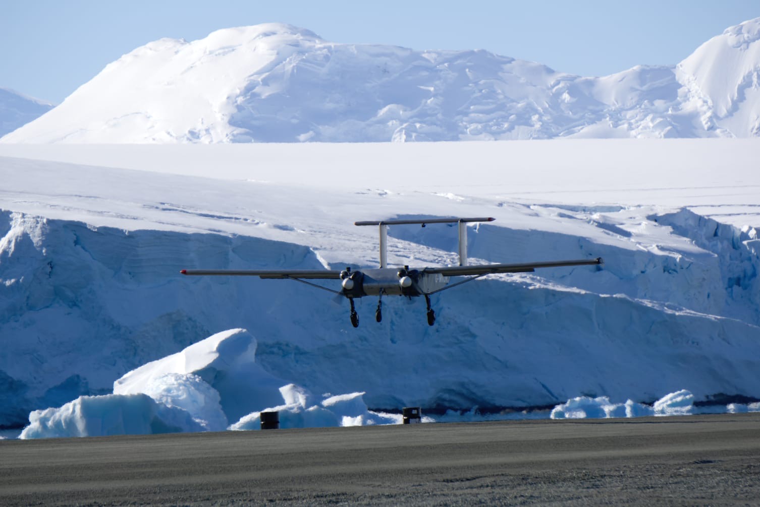 a small, twinengine propeller plane is landing on a runway in antarctica. the plane is in the foreground, with snowcovered mountains and glaciers in the background. the sky is blue and the ice is white.