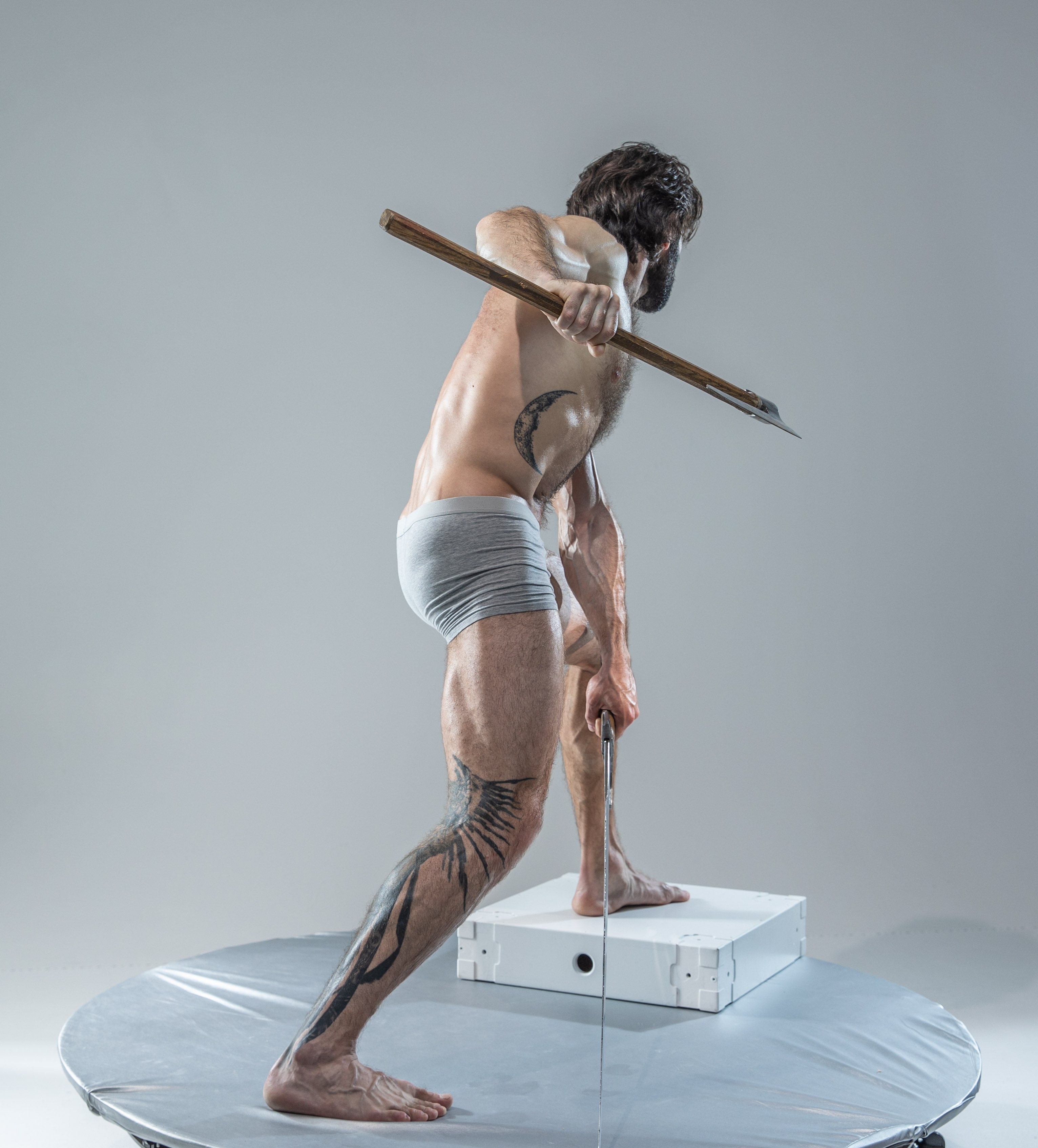 the image features a muscular, tattooed man with a beard, captured in a dynamic pose. he stands on a white platform against a minimalist gray studio backdrop. he holds a wooden axe with a focused expression, suggesting an imminent strike. his physique, tattoos, and the weapon create a powerful and potentially mythological aura. the image uses bright, artificial lighting, highlighting the man's muscular form and the axe's wooden texture. the perspective is from the rear, offering a threequarter view of the man's body. the image evokes themes of strength, action, and potential aggression.