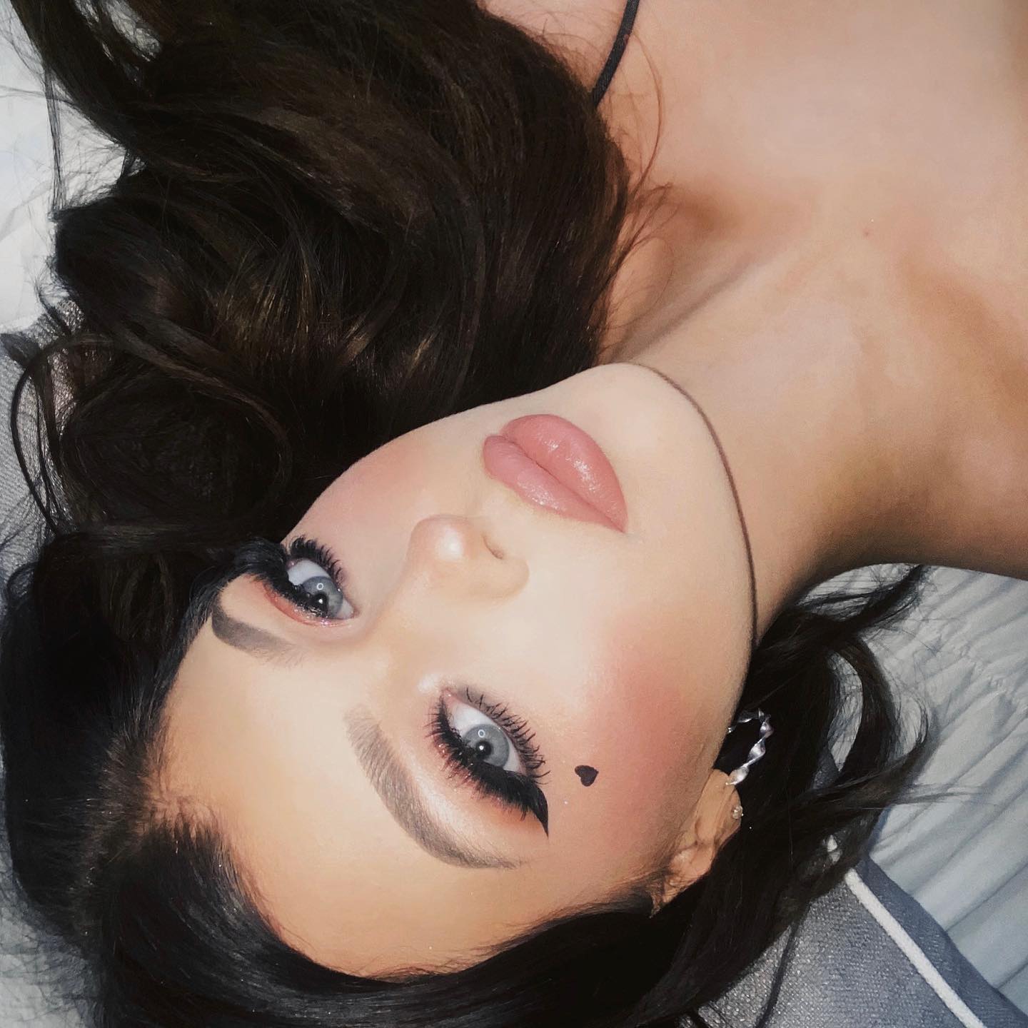 a closeup portrait shows a woman lying on her back on a bed. she is wearing heavy makeup, including winged eyeliner, eyeshadow, blush, and lipstick. a small black heart is drawn on her cheek. her hair is dark and styled around her face. she is wearing earrings. her blue eyes are looking upwards. the shot is taken at an angle, and the background is blurred.