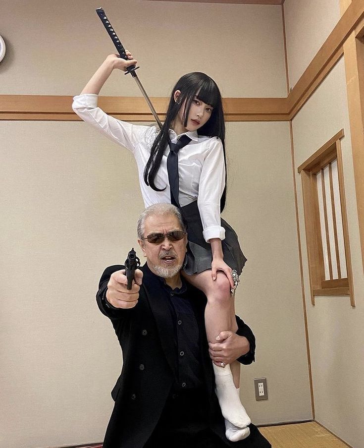 a woman in a school girl uniform holds a sword and sits on a man's shoulders. the man is holding a gun, and both are staring intensely at the camera. they appear to be in a studio, with a wooden wall and floor as the backdrop. the woman is wearing a white collared shirt with a black tie, a black skirt and white socks. the man is wearing a black blazer, a black shirt, and sunglasses.