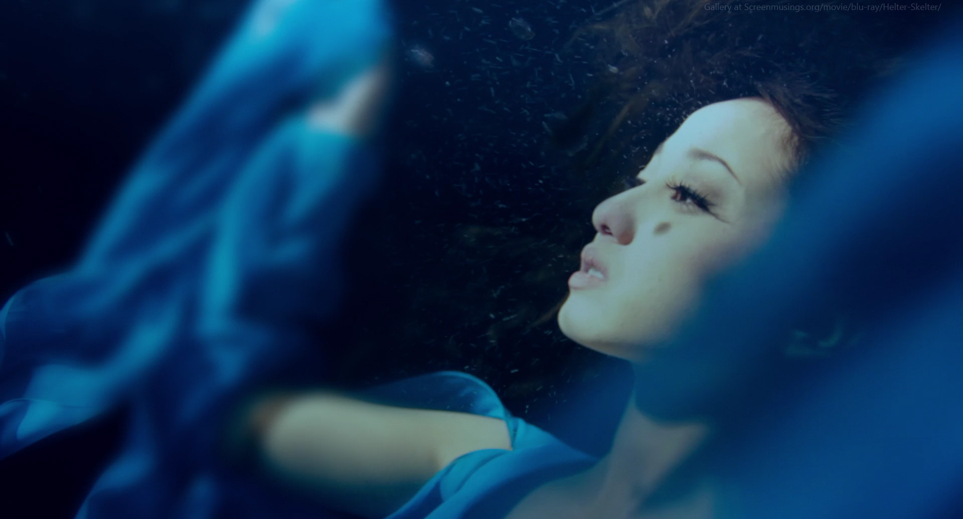 a woman in a flowing blue dress is submerged in dark water. her eyes are open and she has a neutral expression on her face. her hair floats around her head and her arms are outstretched.  the image has a soft, ethereal quality due to the lighting and the way the water distorts the light.