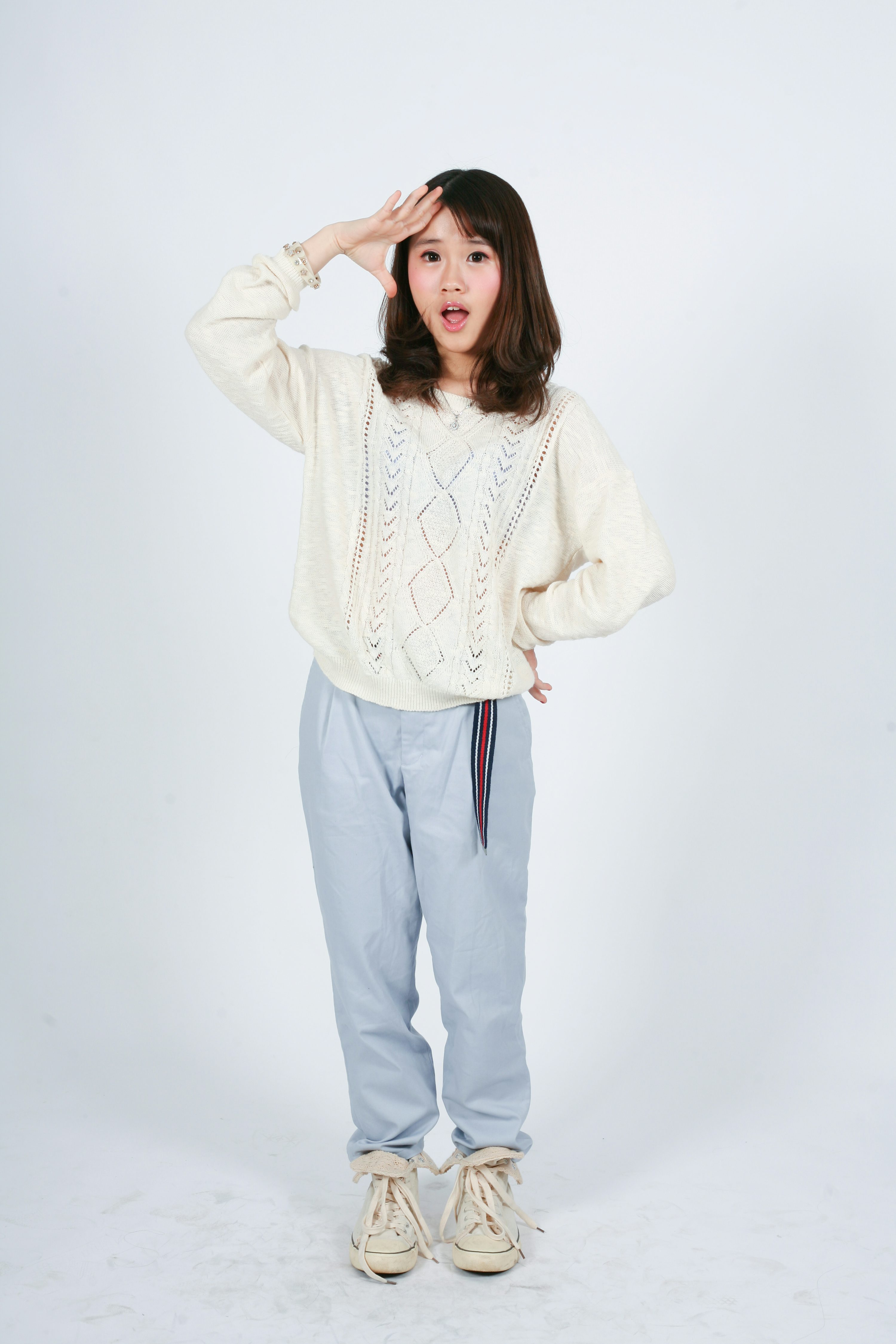 a fullbody shot of a young asian woman in a casual outfit against a white background. she's wearing a creamcolored knitted sweater, light blue pants, and offwhite sneakers.  her hand is on her forehead, and she has a surprised or shocked expression on her face. the image appears to be a studio portrait.