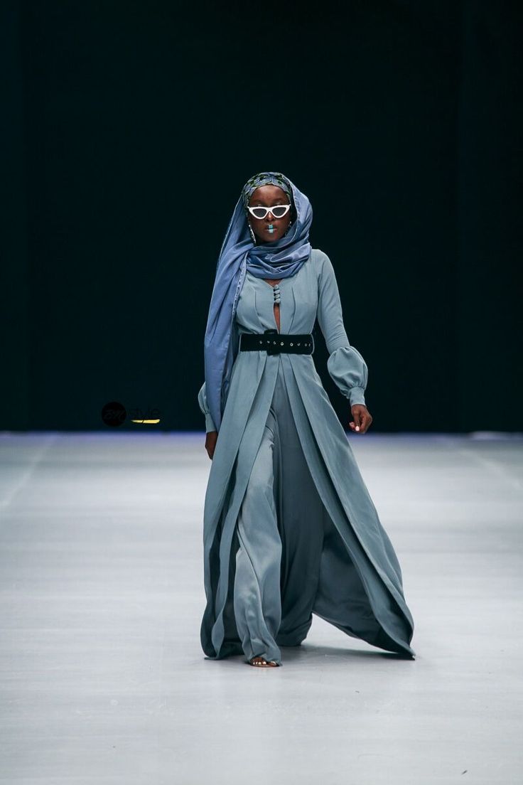 a slender model with dark skin walks down a runway, wearing a long, flowing teal jumpsuit that is styled like a dress. the jumpsuit has a belted waist and wide sleeves. the model wears a matching teal hijab, white cateye sunglasses, and has light makeup. the background is dark and blurry, and the focus is sharply on the model. the overall image is of high fashion, and the style is elegant and sophisticated. the model's confident pose suggests a powerful and stylish look.