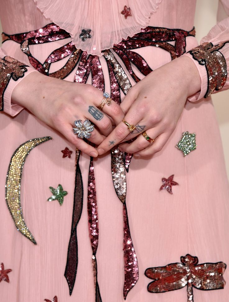 a closeup shot focuses on the hands and lower bodice of a person wearing a pale pink, longsleeved gown. the dress features intricate embroidery with sequins forming various shapes like stars, moons, and dragonflies.  dark pink/maroon ribbons are tied in a bow at the waist. the person's hands are adorned with several gold rings, including a notable floral ring and a ring shaped like a birdcage.  small tattoos are visible on their fingers. the overall style is elegant and highfashion, suggestive of a redcarpet event or similar formal occasion. the dress appears to be of high quality, likely a designer piece.