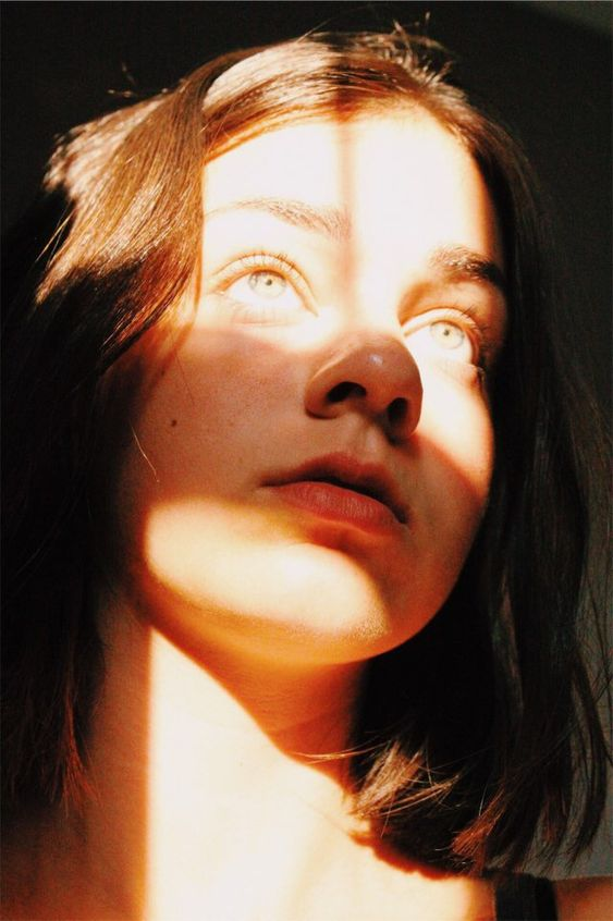 a closeup portrait of a young woman illuminated by sunlight streaming through a window. her face is half in shadow, creating a dramatic contrast. she is gazing upwards, her expression a mix of wonder and contemplation. the worm'seye view adds to the intimacy and intensity of the image.
