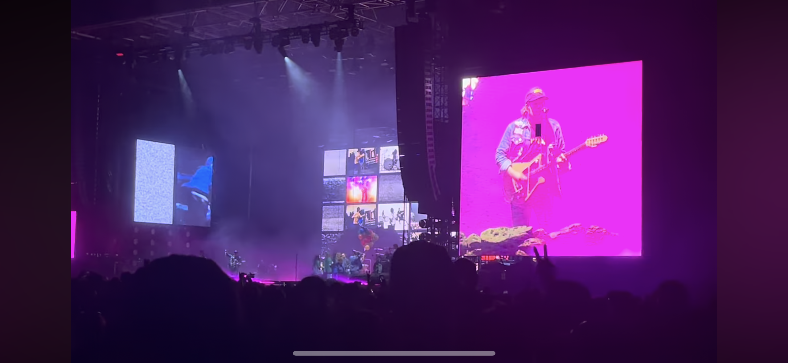 a concert stage with a large video screen displaying a musician playing guitar. the crowd is silhouetted in front of the screen, with the bright lights of the stage and the energy of the crowd visible.