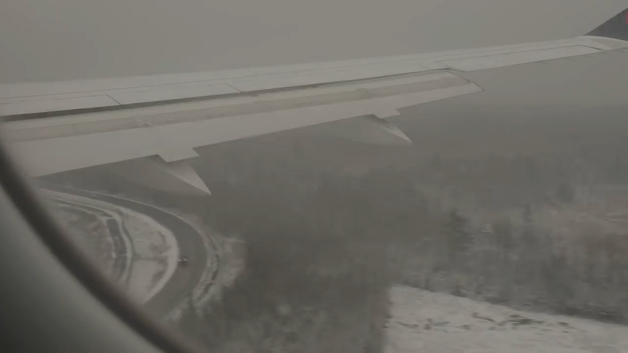 an aerial view from an airplane window showing a snowcovered landscape below. the airplane wing is visible, along with a winding road and a dense forest covered in snow. the sky is overcast, creating a cloudy and diffused light.