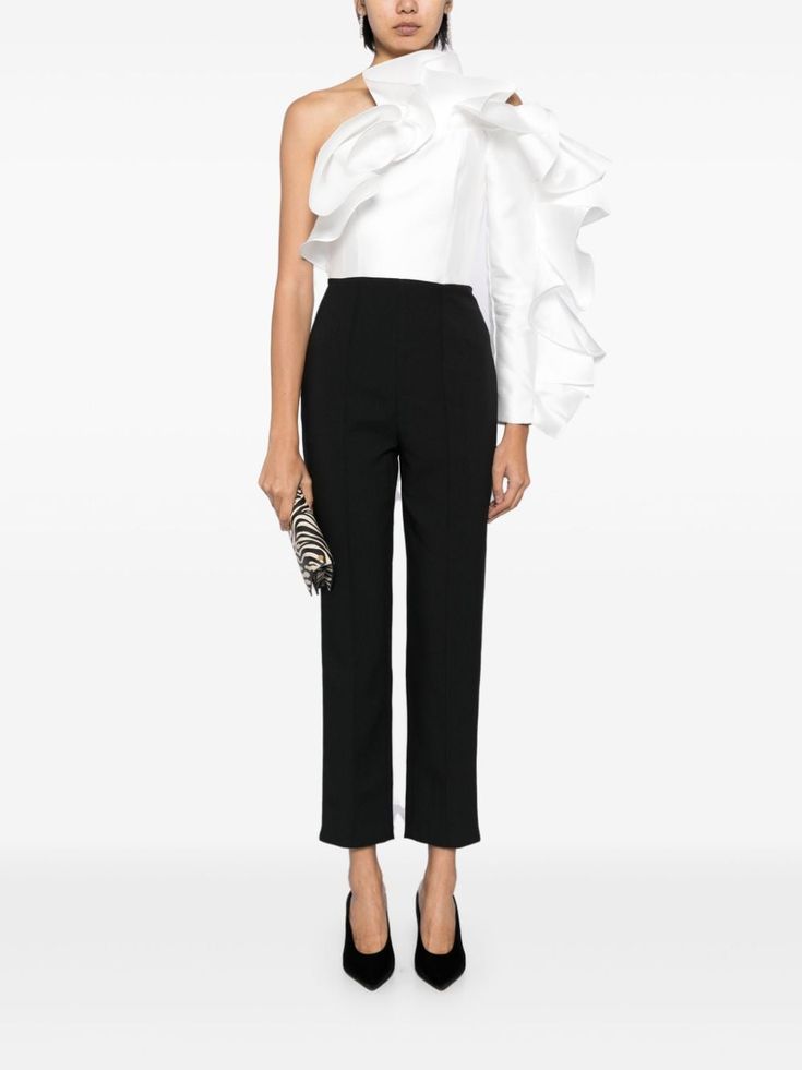 a woman is standing in a studio setting, wearing a white top with a large ruffle on one sleeve. she's wearing black pants and black heels. she's holding a small zebraprint clutch bag in her hand.