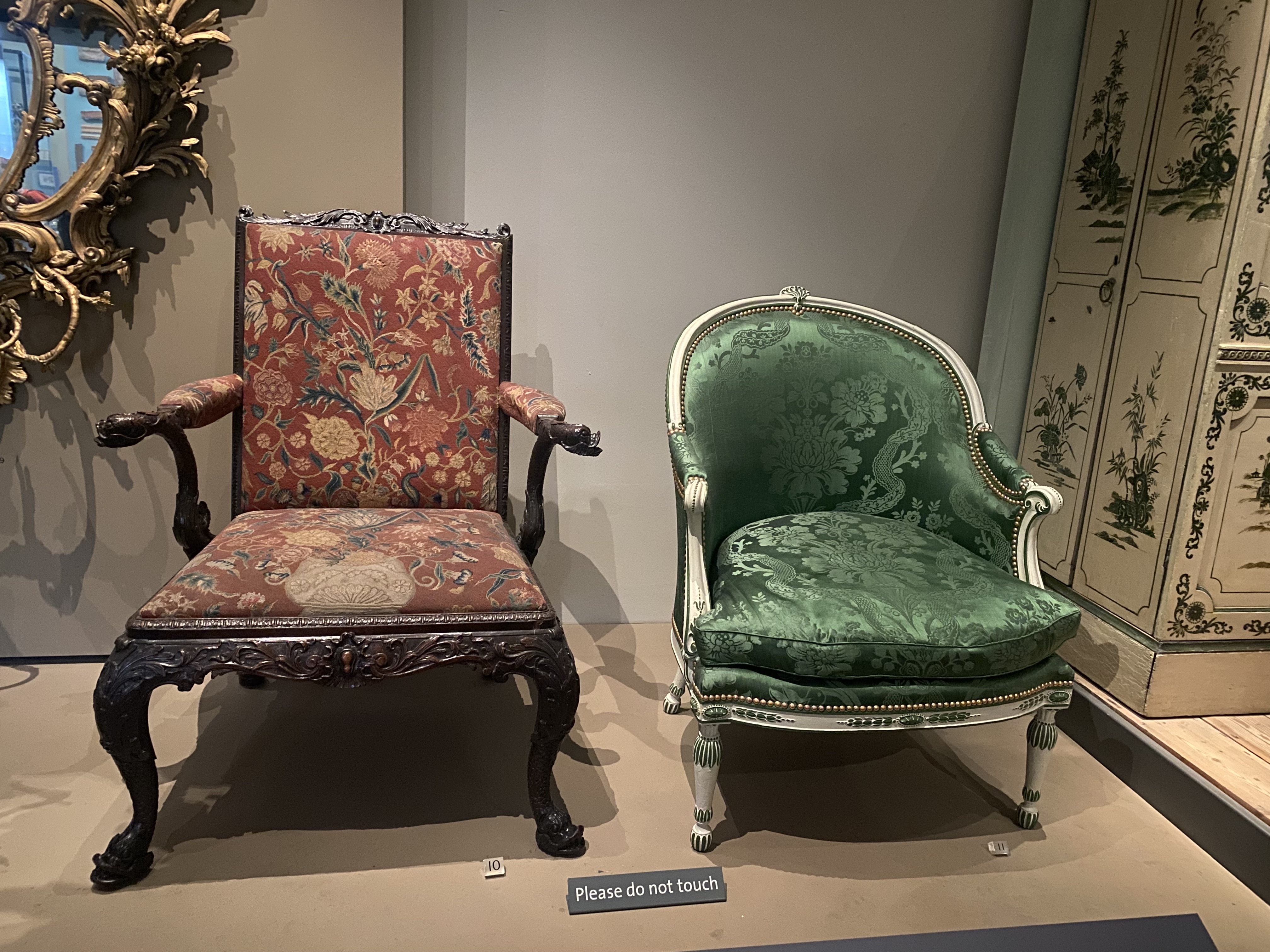 the image shows two antique chairs displayed in a museum setting. the chair on the left has a dark wood frame with carved details and is upholstered in a red fabric with a floral pattern. the chair on the right has a white frame with gold accents and is upholstered in a green fabric with a floral pattern. a gold framed mirror is partially visible on the left, and a decorated cabinet is on the right. a sign that reads "please do not touch" is in the foreground.