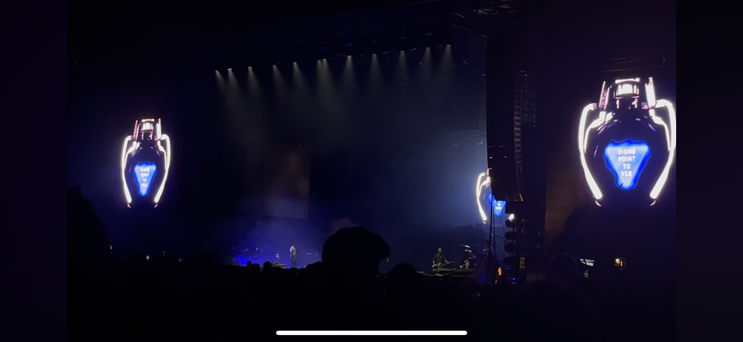 a concert stage with two large glowing heartshaped props, the props have a  white neon outline and a blue interior.  the righthand prop has the text "signs point to yes" inside. the stage has lights shining down on it.  the stage is dark and filled with fog. there is a crowd of people in the audience. a lone silhouette of a singer/musician stands on the stage.