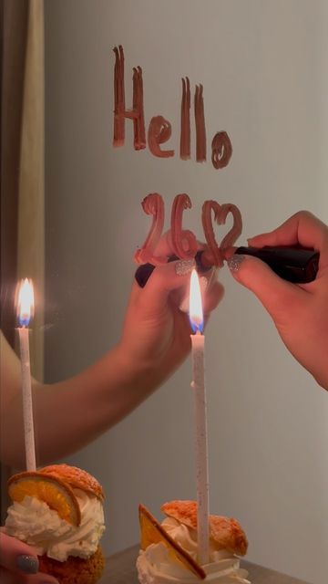 a hand is writing "hello 2023" on a mirror with two lit candles in front of two small cakes with whipped cream on top. the candles are in front of the cakes. the hand is holding a black marker, which is not clearly visible in the photo. there is a red love heart between the 2 and 3. the cakes are slightly blurred in the background.