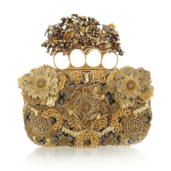 the image shows a goldcolored, ornate clutch bag. the bag is heavily decorated with beads, floral designs, and what appears to be a gold knuckle duster as a handle. the overall design is intricate and eyecatching, suggesting a luxury or designer item. it appears to be a fashion accessory, likely an evening bag or clutch. the clutch is resting on a white surface with a slight reflection below it.