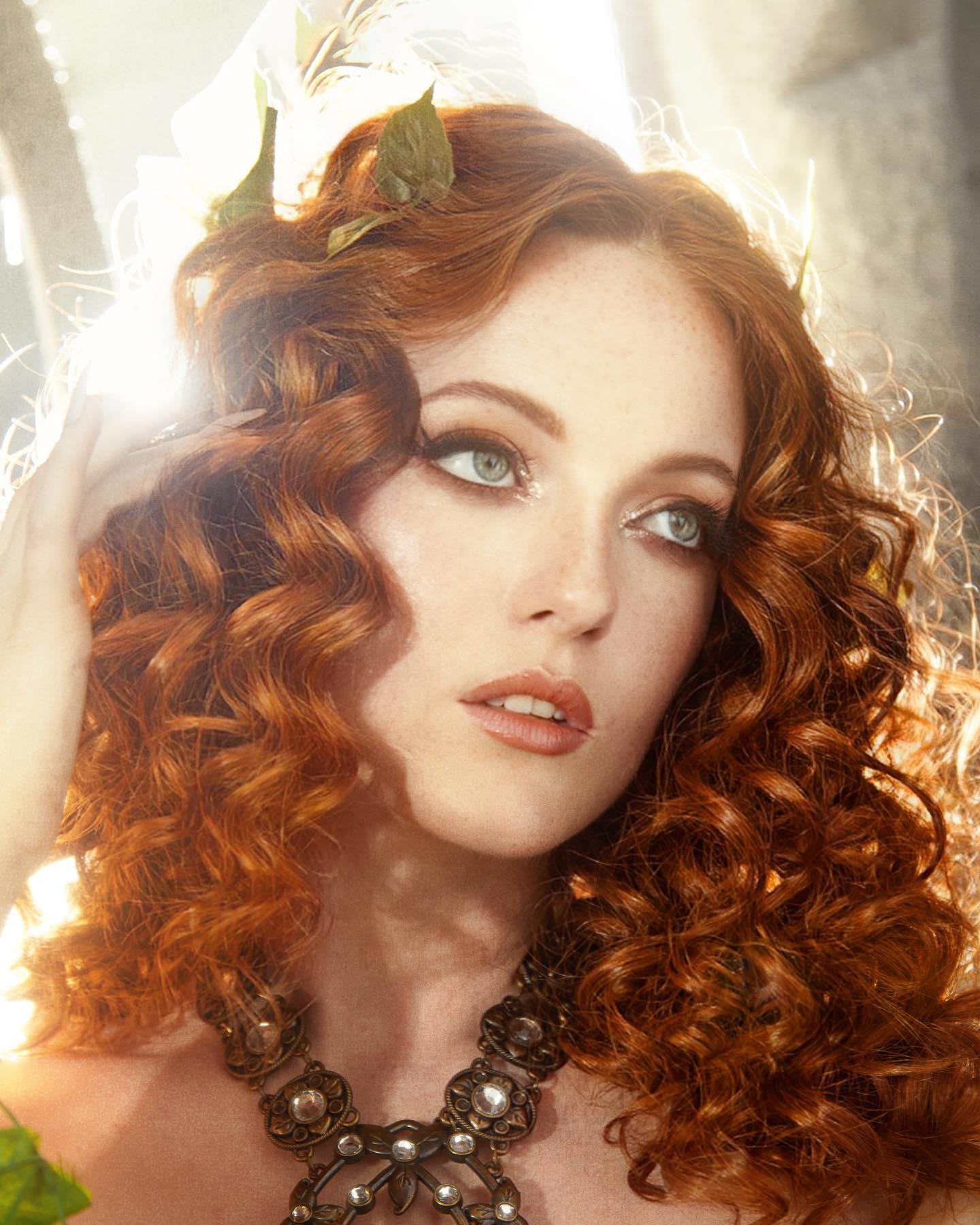 a stunning redhead woman with captivating green eyes is the focal point of this portrait. her fiery, cascading curls frame a face adorned with delicate freckles. she wears a statement necklace featuring intricate metalwork and shimmering stones, adding a touch of glamour to her ethereal look.  a soft, diffused light bathes her features, highlighting the natural beauty of her complexion and the vibrant hue of her hair. leaves, reminiscent of a woodland nymph, are gently tucked into her curls.