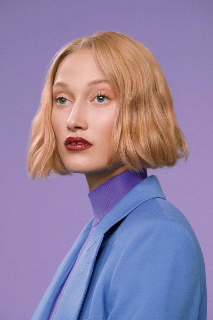 closeup portrait of a young woman with a stylish blonde bob haircut. she's wearing a light blue blazer over a purple turtleneck.  her makeup includes red lipstick. the background is a muted purple. the overall image is sophisticated and fashionable.