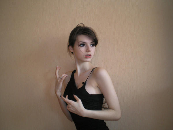 the image shows a young, slender woman with dark brown hair styled in a loose updo. she's wearing a simple, black, sleeveless dress and is posing against a light beige wall. her expression is somewhat pensive or thoughtful, and she is looking away from the camera. her hands are gesturing near her body. the overall aesthetic is elegant and somewhat casual.  the lighting and background are simple, focusing attention on the woman.