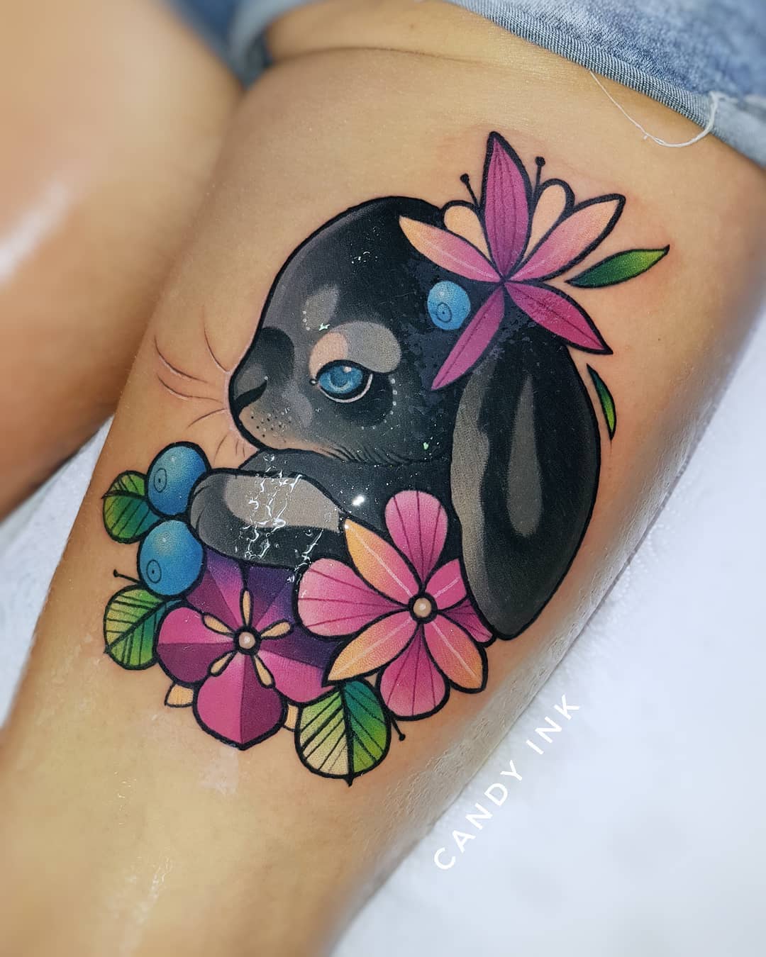 a closeup shot showcasing a freshly done neotraditional tattoo of a grey bunny nestled amongst colorful flowers and blueberries on a woman's thigh. the tattoo is executed in a vibrant color palette and boasts clean lines.