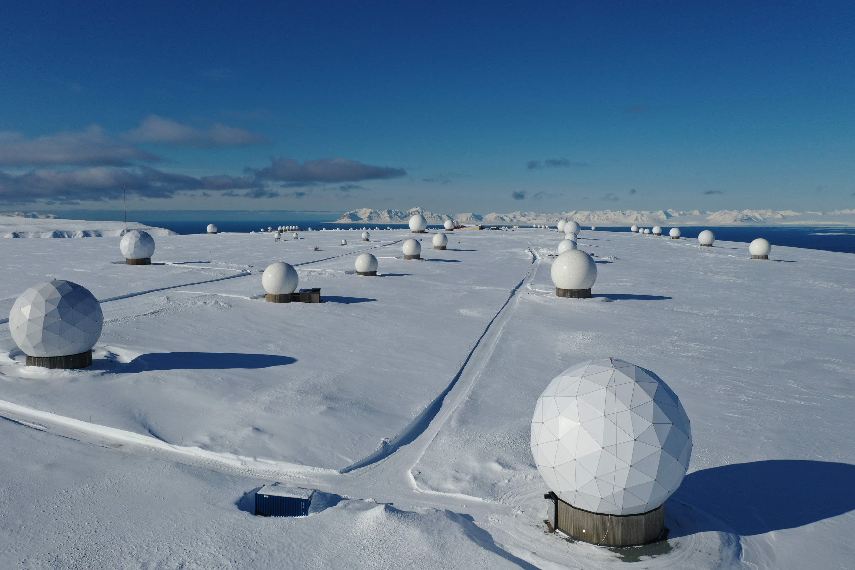 an aerial view of a snowy landscape with a line of radar antennas stretching across the horizon. the antennas are white and dome shaped, and they are set against a backdrop of snowcovered mountains and a blue sky. the photo was likely taken in a remote location near the arctic circle.