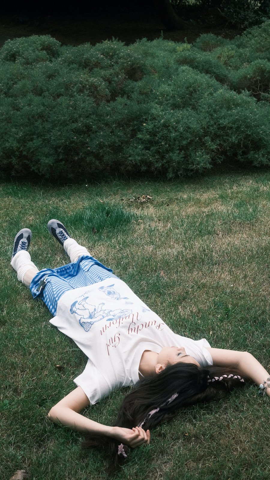 a young woman with long dark hair lies on her back in a grassy park. she's wearing a white tshirt with a design, a blue and white checkered skirt, and white socks with grey sneakers. her eyes are closed and she appears relaxed and peaceful. the scene is serene and idyllic, suggesting a summer afternoon.
