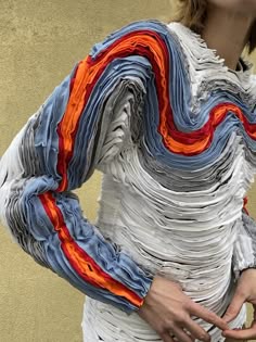 a woman is wearing a white, blue and orange layered garment. the design of the garment has a unique and abstract feel, with the fabric being pleated and folded into complex, swirling shapes. the garment appears to be made of many different layers of fabric, with the blue and orange layers creating a striking contrast against the white base. the garment is being worn by a woman who is standing in a studio setting. the woman's hands are visible in the image, and they are holding the fabric of the garment. the woman's face is partially visible, and she is looking at the camera. the background of the image is a plain, neutral color. the overall effect of the image is one of creativity, fashion, and art. the woman appears to be wearing a sculpture as a garment.
