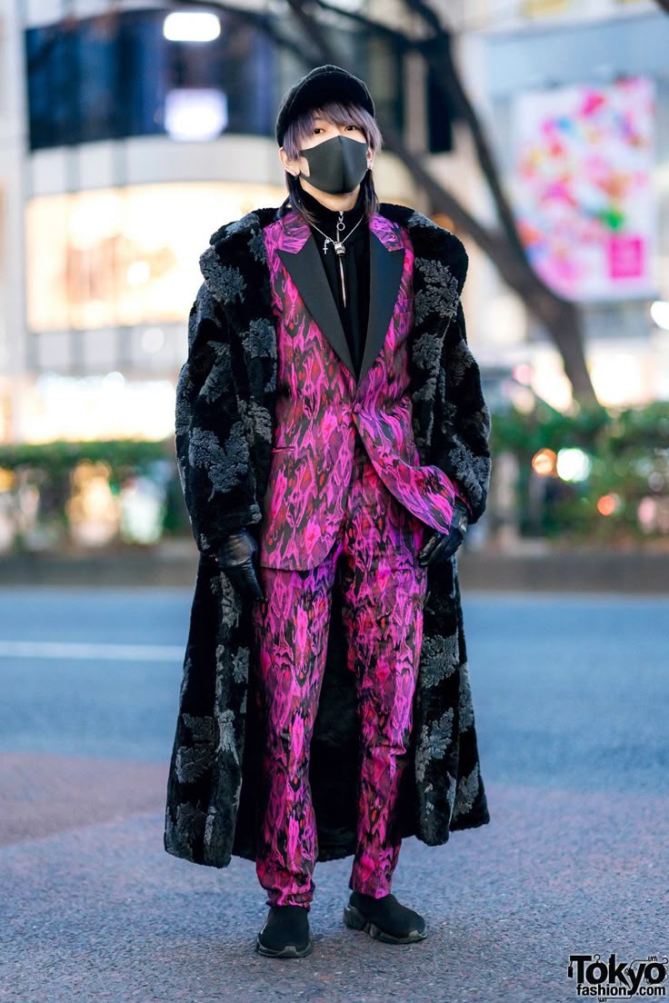 a person stands outside in the street wearing a hat, mask, long coat, suit, gloves, and shoes. the coat is long and black with a flower pattern and reaches almost to the ground. the suit is a colorful pink and purple pattern with black lapels and a black shirt underneath. the person also has a cross necklace on and is wearing black gloves.