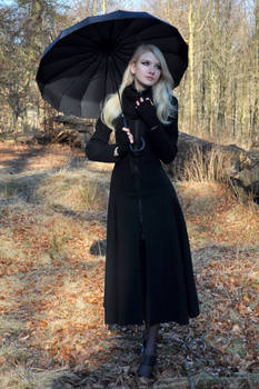 a blonde woman in a long black coat and gloves stands in a forest holding a black umbrella.  the autumn leaves are on the ground around her.  she is dressed in a gothic style.  the image has a dark and mysterious feel.