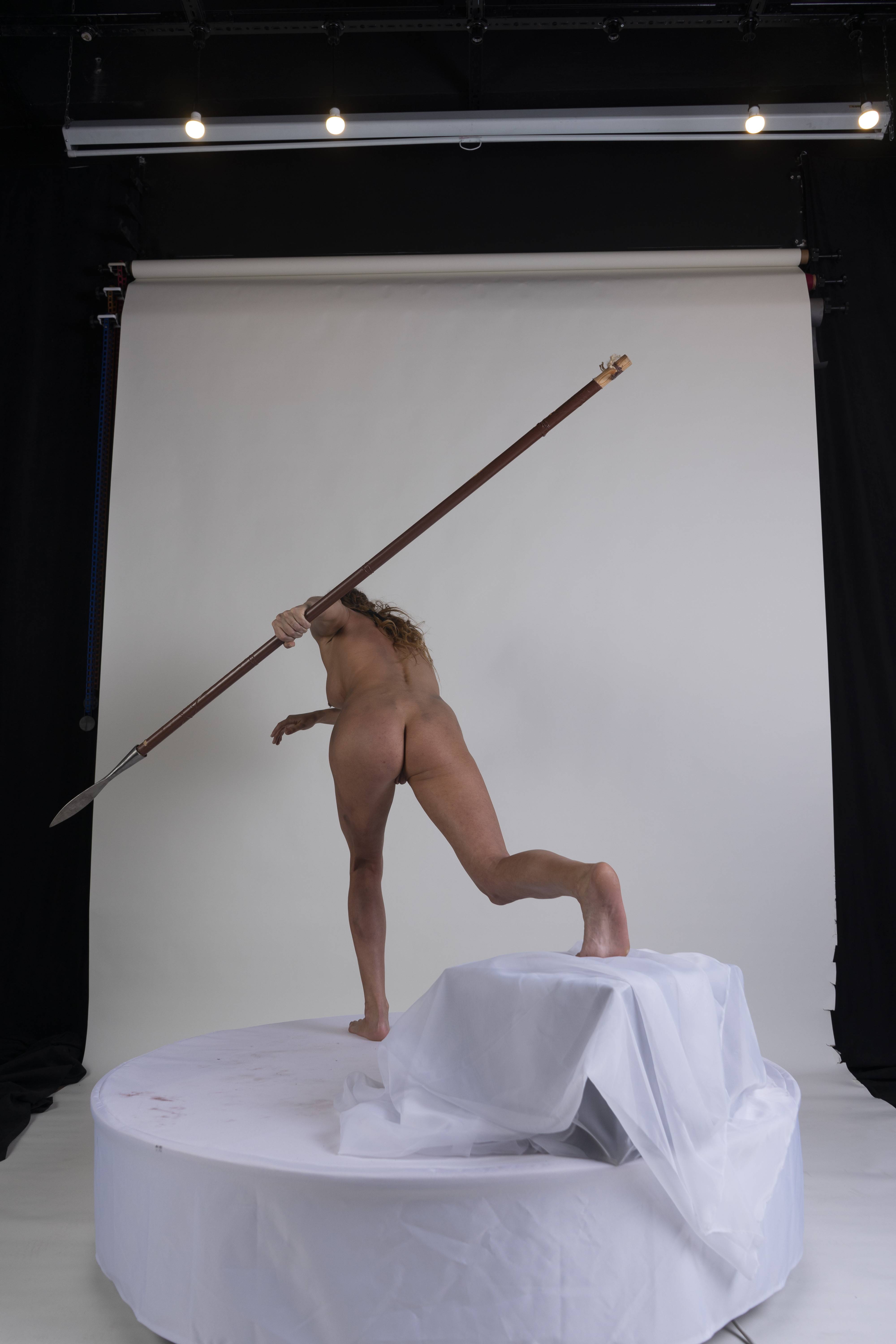 a nude woman stands on a stage with a backdrop, holding a spear.